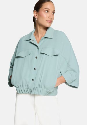 Femme portant une veste oversize bleu clair avec boutons-pression et ourlet élastique, associée à un pantalon blanc, les mains dans les poches, regardant vers la droite.