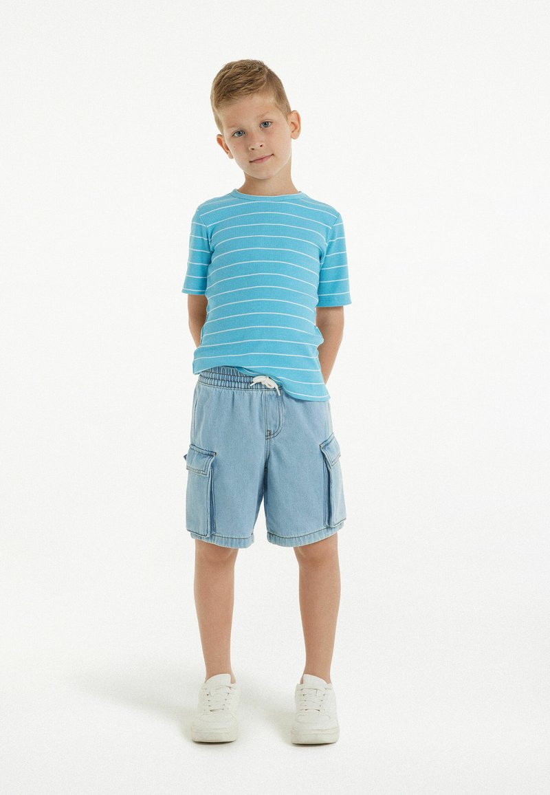 Chemise à rayures bleu clair à manches courtes associée à un short cargo en denim. Les shorts ont des poches sur les côtés et une taille élastique. Baskets blanches.