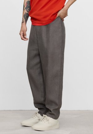 Persoon die losse grijze broek, witte sneakers en een rood shirt met zichtbare tatoeages op één arm draagt tegen een effen achtergrond.