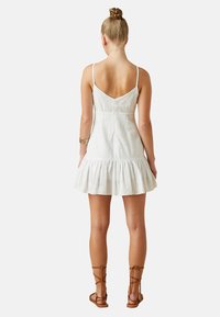 Vestido mini blanco con textura y dobladillo acampanado, tirantes ajustables en forma de espagueti y espalda en forma de V. Combinado con sandalias marrones con tiras.