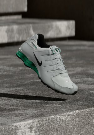 Chaussure de course Nike blanche et noire avec une semelle intercalaire amortissante verte en suspension au-dessus d'une surface en béton et des marches en arrière-plan.