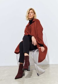 Cappotto color ruggine con bottoni, indossato sopra un dolcevita nero e abbinato a stivaletti alla caviglia bordeaux. Sedia trasparente su un pavimento chiaro.