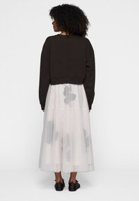 Brun cropped sweatshirt båret over en hvid tyl nederdel med grå blomster mønstre. Sorte sko fuldender outfittet. Ensfarvet baggrund.
