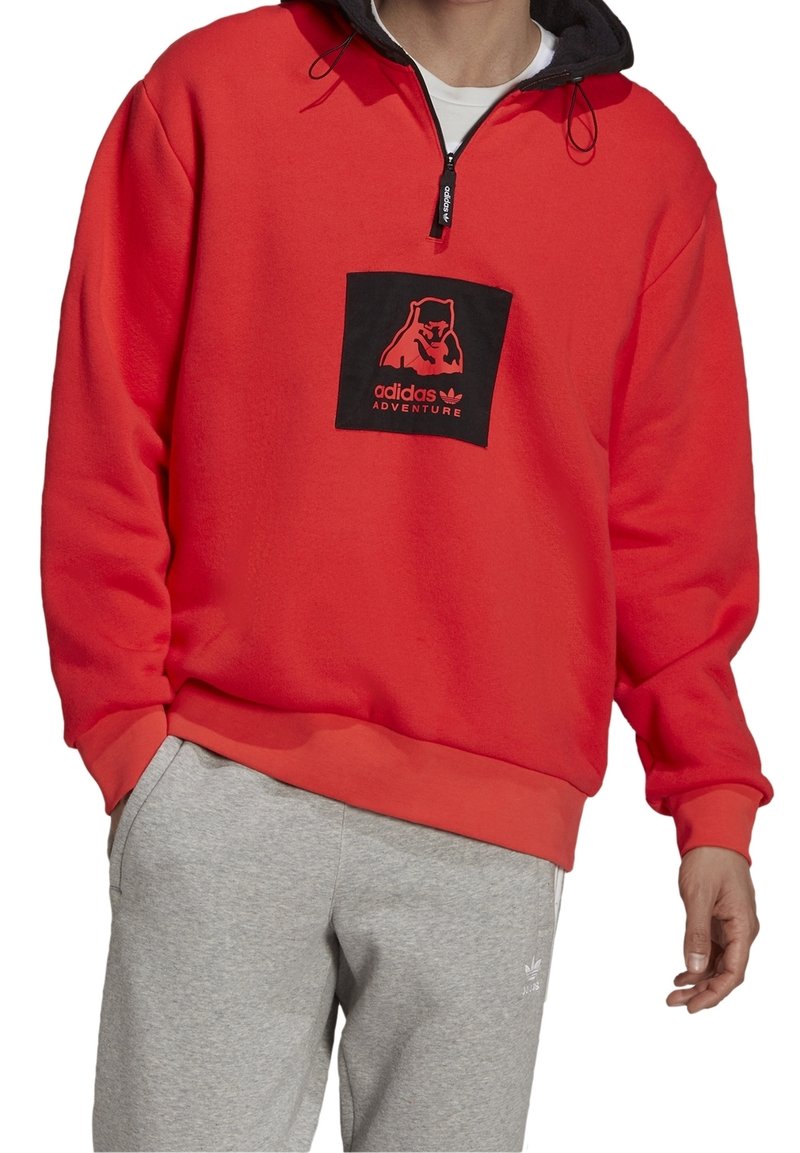 Pullover in pile rosso con una toppa quadrata nera raffigurante un orso e la scritta "adidas ADVENTURE"; dotato di colletto con zip e dettagli grigi.