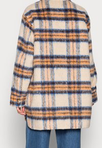 Fleecejacka med en ljus krämfärgad bas, prydd med ett rutmönster i orange, blått och brunt. Inkluderar knappar vid ärmsluten.