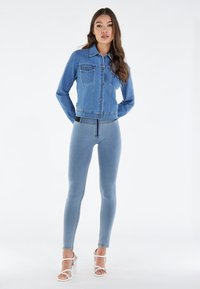 Nainen seisoo valkoisella taustalla pukeutuneena istuvaan siniseen farkkutakkiin, vaaleansinisiin korkeavyötäröisiin kapealahkeisiin farkkuihin ja valkoisiin korollisiin sandaaleihin.