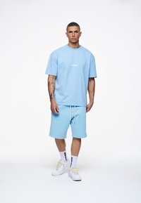 Lyseblå bomullst-skjorte med rund hals og logo. Kombinert med matchende lyseblå shorts med snøring og en avslappet passform. Hvite sneakers.