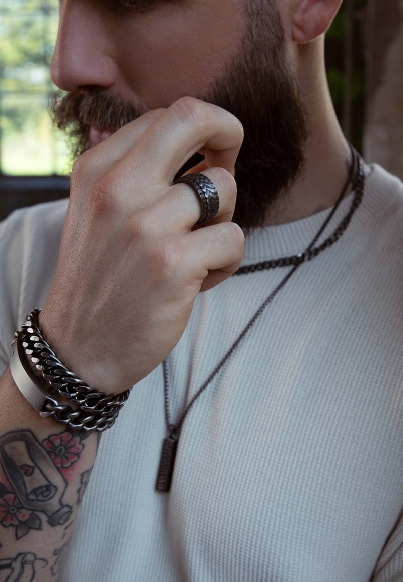 Zwarte metalen ring en gelaagde kettingen op een hand, die textuurmaterialen laten zien. De outfit bestaat uit een geribd, lichtgekleurde shirt.
