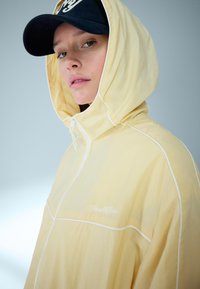 Mlada oseba v svetlo rumeni jakni s kapuco in belim trakom ter črno baseball kapo, rahlo obrnjena v stran na enobarvni podlagi.