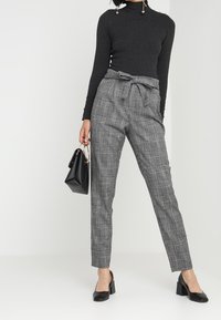 Černý žebrovaný rolák s dlouhými rukávy, kombinovaný s šedými kostkovanými kalhotami s vysokým pasem a mašlí v pase, a černými botami na blokovém podpatku.