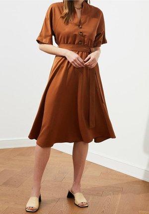 Vestido marrón de largo hasta la rodilla, con mangas cortas, botones en la parte delantera y un cinturón a juego. Tela suave con una silueta fluida.