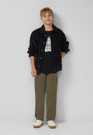 Schwarze Cordjacke mit Knopfleiste, getragen über einem schwarzen T-Shirt mit Grafik. Olivgrüne Hose und weiße Sneakers vervollständigen den Look.