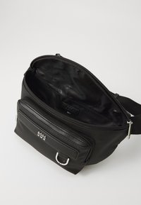 Black nylon crossbody bag with zippered compartments, featuring a front pocket and metallic hardware accents. Interior is dark with branding.