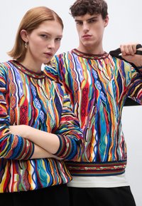 Pulls colorés en tricot avec des motifs complexes en bleu, rouge, jaune et vert. Poignets et col côtelés, présentant des motifs géométriques.