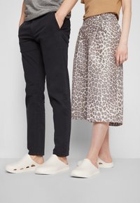 Homme et femme debout côte à côte portant des sandales blanches à enfiler, l'homme en pantalon noir et chemise grise, la femme en jupe imprimé léopard et haut beige.