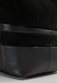 Czarna zamszowa torba z gładką skórzaną podstawą. Charakteryzuje się teksturowaną powierzchnią, płaskim kształtem i kontrastującym skórzanym paskiem w środkowej części.