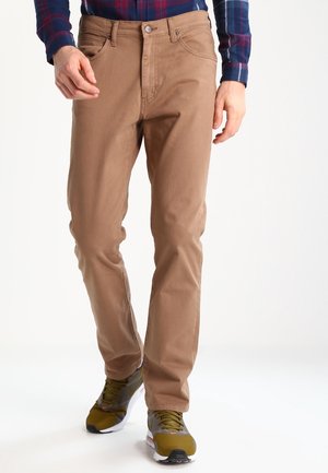 Mann trägt beige gerade geschnittene Hose, grün-graue Sneaker und ein blau-rotes kariertes Hemd, läuft vor weißem Hintergrund.