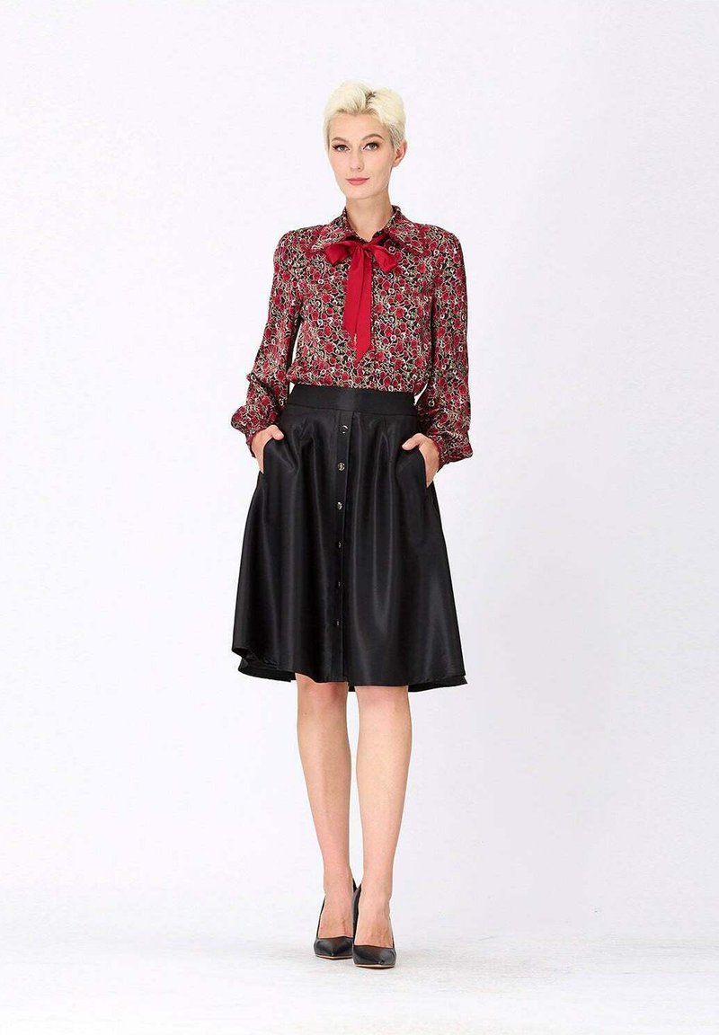Blouse florale rouge avec un nœud au cou, associée à une jupe noire en satin arrivant aux genoux avec un design à boutons sur le devant et des poches latérales. Talons noirs.