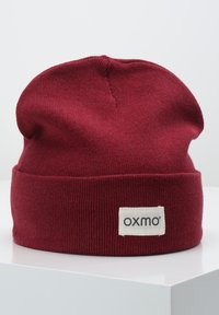 Red knitted beanie with a folded cuff, featuring a woven label with the brand name "oxmo." Soft texture and slouchy design.