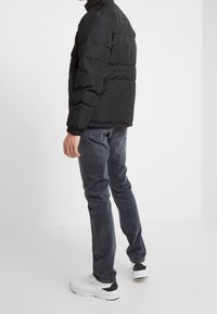 Doudoune noire avec col montant et poignets élastiques, associée à un jean foncé et des baskets blanches, vue de dos.