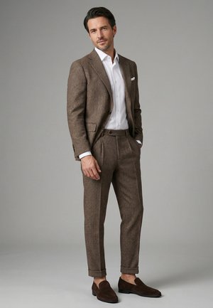 Mand stående med afslappede hænder iført brun sildebenet jakkesæt, hvid skjorte og brune loafers mod grå baggrund.