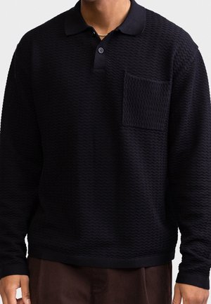 Homme portant un polo noir texturé à manches longues avec une poche poitrine et un pantalon marron, vu des épaules à la taille.