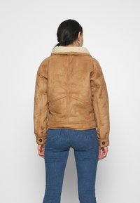 Brun mocka jacka med en krämfärgad shearling-krage, har en åtsittande design, långa ärmar och knäppte ärmslut. Bärs med blå jeans.