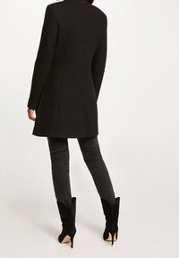 Zwarte wollen jas met een getailleerde snit, lange mouwen en een splitdetail aan de achterkant, gecombineerd met slim-fit zwarte broek en puntige laarzen.