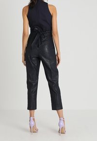 Černý top bez rukávů s vysokým límcem, spárovaný se lesklými černými kalhotami s vysokým pasem a fialovými sandály na podpatku. Viditelný zadní pohled.