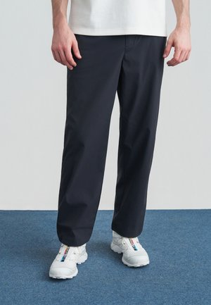 Muž nosiaci voľné čierne nohavice a biele tenisky s viacfarebnými pruhmi, stojaci na modrom kobercovom povrchu pred bielou stenou.