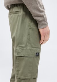 Άτομο που φοράει ελαιόχρωμο παντελόνι cargo με μεγάλη πλαϊνή τσέπη και ένα μανίκι από πλεκτή μπλούζα σε χρώμα ταούπ ορατό κοντά στο χέρι.