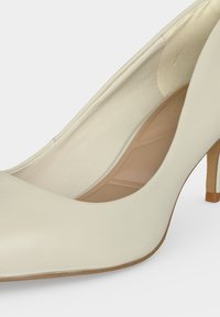 Escarpin beige à talon haut, bout pointu, extérieur lisse, semelle intérieure rembourrée et talon fin sur fond blanc.