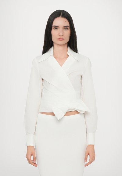Blouse blanche à manches longues avec un col et un accent nœud à l'avant. Tissu doux et léger ; design ajusté. Associée à une jupes blanche en maille côtelée.