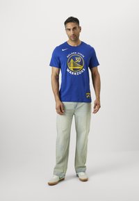 Tricou din bumbac albastru cu logo-ul "Golden State Warriors" în galben, asociat cu blugi deschiși la culoare și adidași albi cu accente verzi.