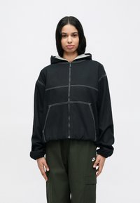 Μαύρο φούτερ με φερμουάρ, με αντιθετικές λευκές ραφές, γκρι εσωτερική κουκούλα και πλαϊνές τσέπες, συνδυασμένο με παντελόνι σε ελιάς πράσινο χρώμα.