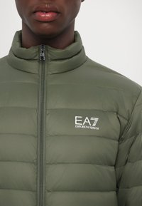 Olive green puffer jacket with a high collar, zip closure, horizontal stitching, and a white EA7 logo on the left chest. Lightweight and textured.