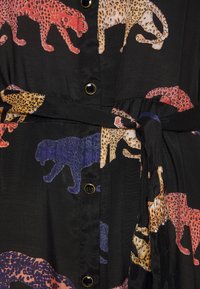 Tissu noir avec un motif léopard coloré en rouge, jaune et bleu, comportant des boutons et une ceinture nouée à la taille.