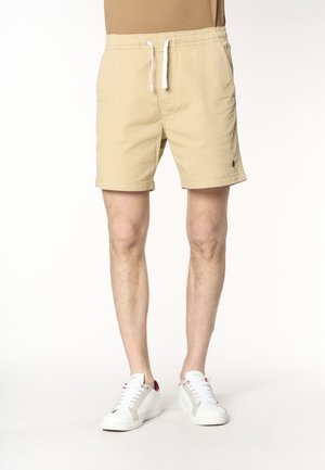Uomo che indossa pantaloncini beige con coulisse e scarpe da ginnastica bianche, in piedi su uno sfondo bianco semplice.
