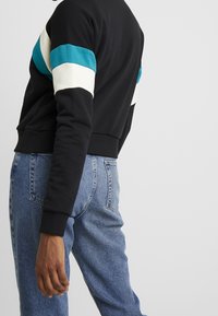 Zwarte sweatshirt met turquoise en witte horizontale strepen op de bovenste mouwen. Gemaakt van zachte stof; voorzien van een geribde zoom en manchetten.