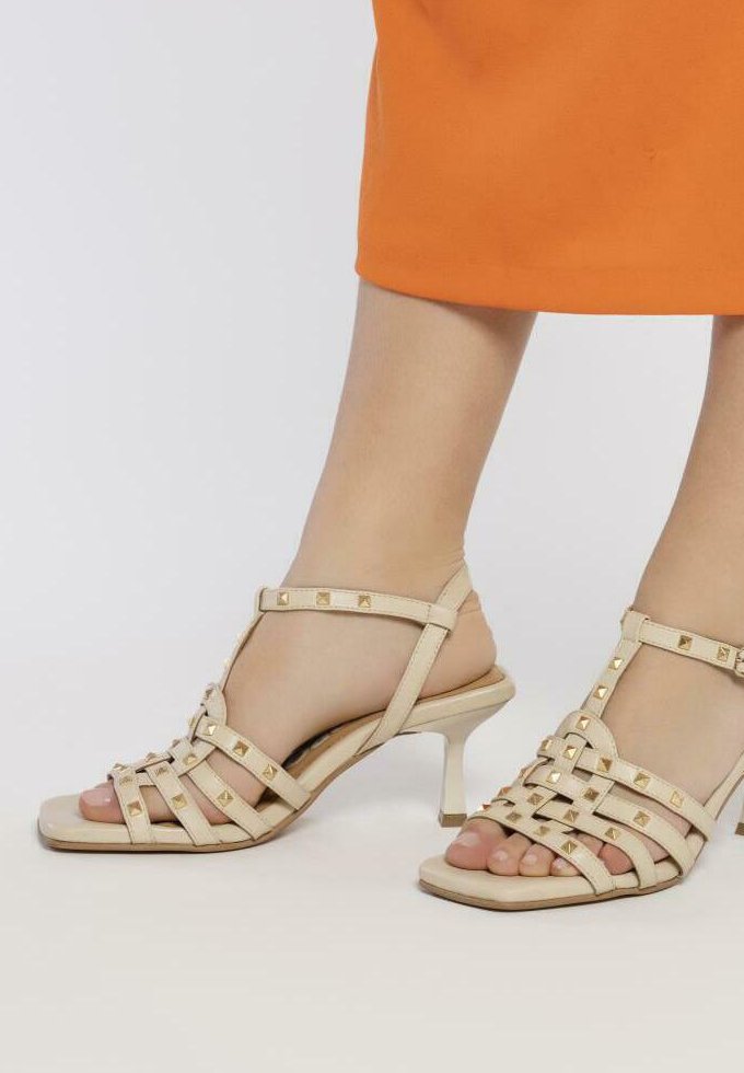 Sandalias de color beige con tiras y un tacón bajo, que presentan acentos de tachuelas doradas, correas texturizadas y un diseño de punta abierta, sobre un fondo naranja.