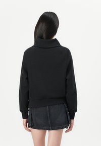 Woman with long black hair facing away, wearing a black long-sleeve sweatshirt and a short dark denim skirt against a plain white background.