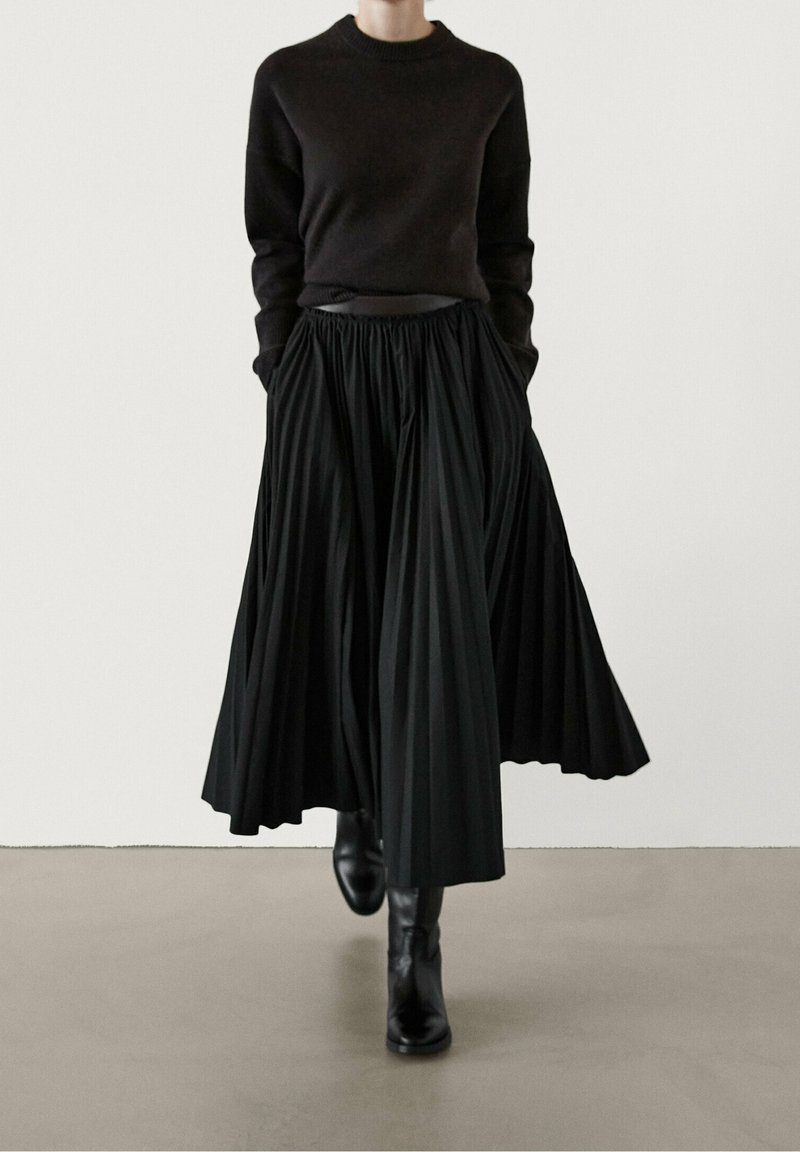Personne portant un pull noir à manches longues et un pantalon noir plissé à jambes larges, associé à des bottines noires, debout devant un fond clair uni.