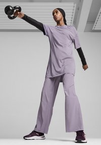 Šviesiai violetinė sportinė apranga, sudaryta iš laisvo marškinėlių ir plačių kelnių, derinama su juodais sportbačiais. Modelis laiko girą.