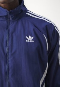 Navy blue Adidas track jacket with a full-length zipper, white three-stripe detail on sleeves, and white logo on the left chest. Smooth texture.