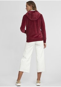 Burgunderfarget, fløyels zip-up hettegenser med hette, kombinert med hvite, korte vide bukser med frynsete kanter, og beige sneakers.