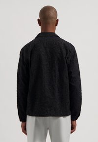 Svart strukturert jakke med preget mønster, lange ermer og krage, kombinert med lysegrå bukser.