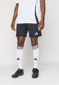 Μαύρο σορτσάκι ποδοσφαίρου με γκρι λεπτομέρειες, λευκές κάλτσες με μαύρες ρίγες και μαύρα παπούτσια ποδοσφαίρου με χρυσές λεπτομέρειες. Λευκή αθλητική μπλούζα.