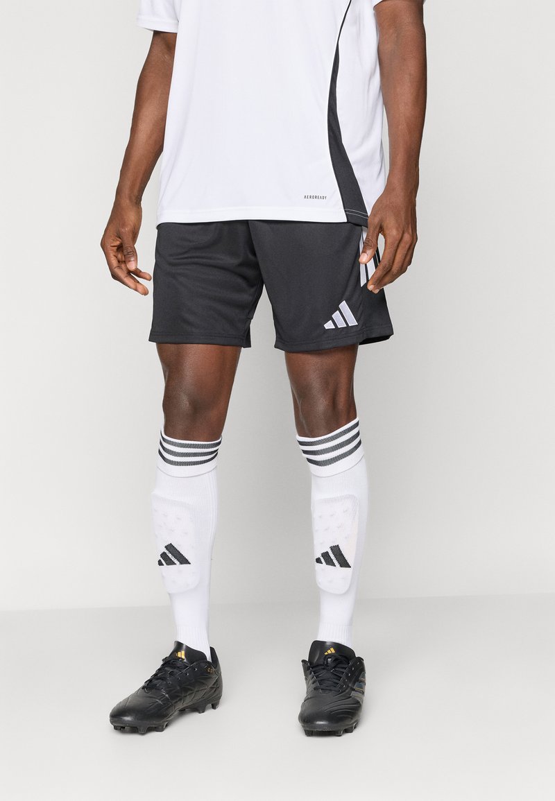 Μαύρο σορτσάκι ποδοσφαίρου με γκρι λεπτομέρειες, λευκές κάλτσες με μαύρες ρίγες και μαύρα παπούτσια ποδοσφαίρου με χρυσές λεπτομέρειες. Λευκή αθλητική μπλούζα.