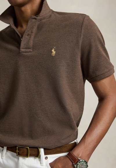 Mees kannab pruuni polosärki kollase tikitud logoga, valgeid pükse, pruuni seemisnahast vööd ja kellaga, millel roheline kellanägu ja pruuni nahast rihmaga.