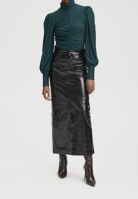 Femme portant un chemisier vert sarcelle froncé à col montant, une jupe midi noire brillante à texture crocodile, et des bottes à talons hauts marron foncé, debout sur un fond blanc.